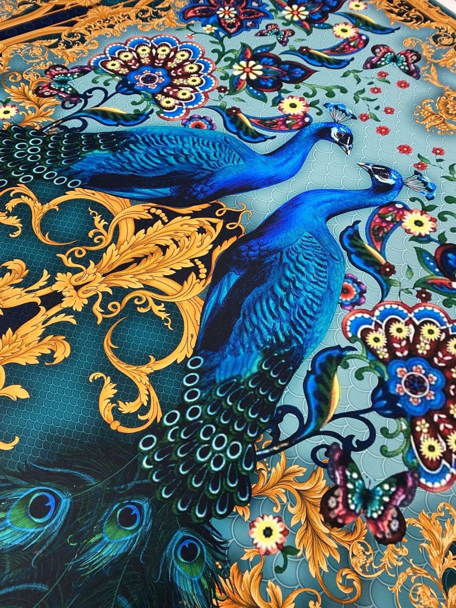 Close-up of vintage peacock pattern fabric with vibrant blue peacocks, gold ornate details, and colorful floral accents on a light blue background